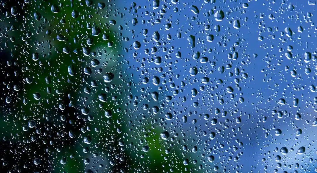 Close-up of a car window with numerous water droplets beading on the surface, with a blurred green and blue background.