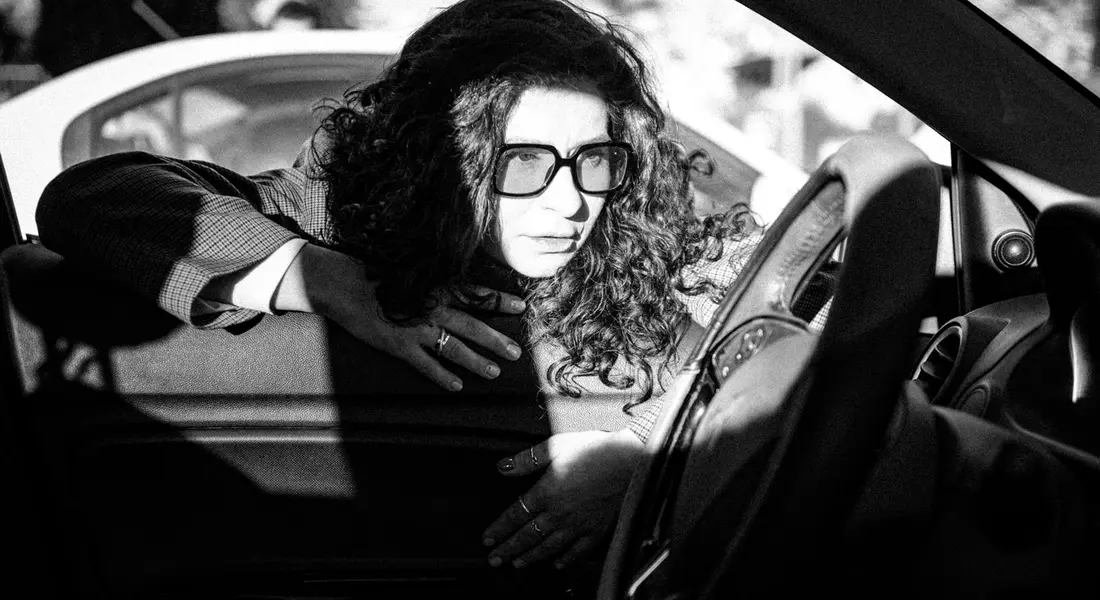 Person wearing glasses sits in a car and looks out from the driver's side window, with a serious expression.