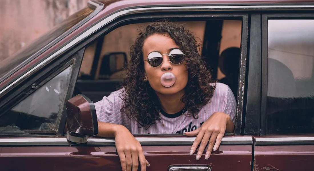 Person with round sunglasses leaning out of a car window and blowing a bubble with gum.