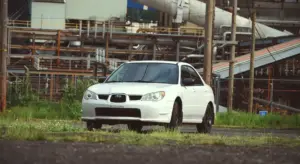 A white SUV parked in an industrial area with pipes and metal structures in the background.