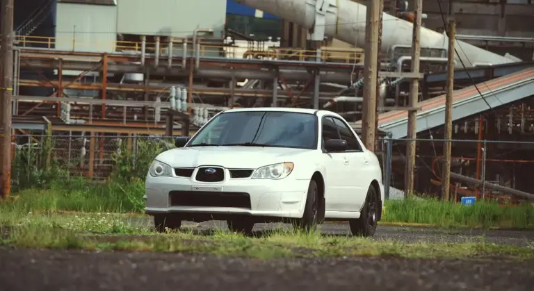 A white SUV parked in an industrial area with pipes and metal structures in the background.