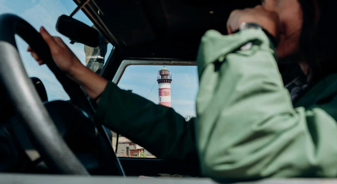 A person wearing a green jacket sits in the driver's seat, reaching toward the windshield, with a lighthouse visible through the front window.