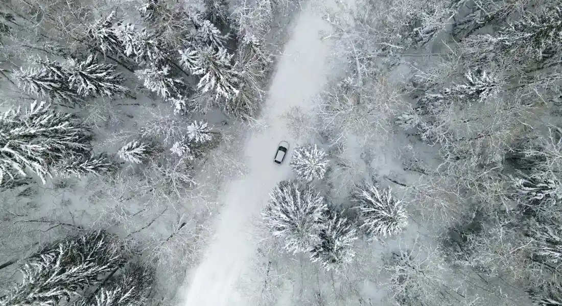 Aerial view of a car driving along a snow-covered forest road, illustrating winter prep for protecting paint.