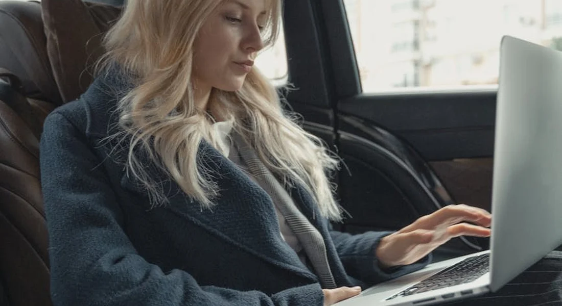 Blonde woman seated in a car interior, working on a laptop placed on her lap.