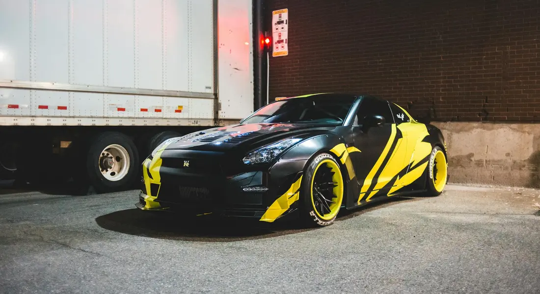 Black sports car with a bright yellow and black vinyl wrap, parked in an urban setting near a loading dock.