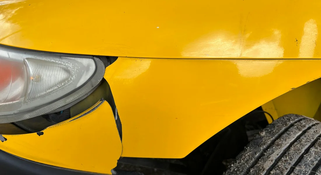 Close-up of a yellow plastic car bumper with visible cracks and damage near the headlight and wheel.
