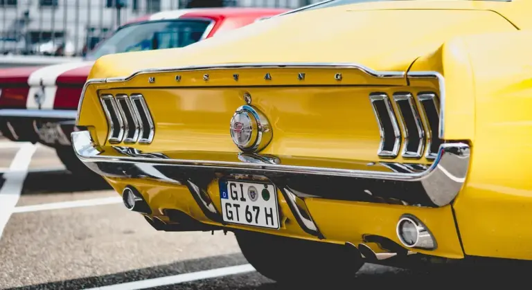 Rear view of a yellow vintage Ford Mustang with chrome bumper and distinctive tri-bar tail lights in a parking lot.